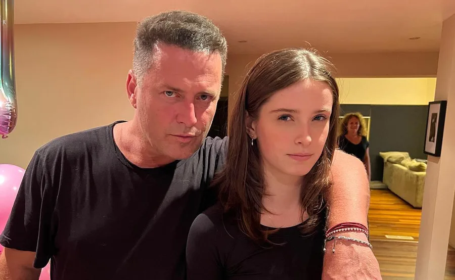 Father and daughter together indoors, with a woman in the background, pink balloons visible to the side.