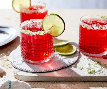 Cranberry margaritas with lime garnish in textured glasses on a sunny countertop.