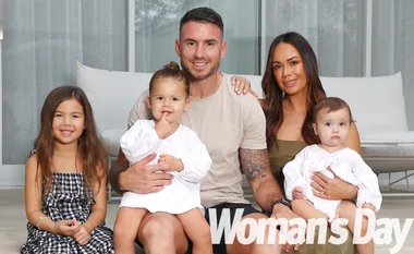 "Family sitting together on a couch, two young children in white outfits, parents smiling. Woman’s Day logo at bottom."