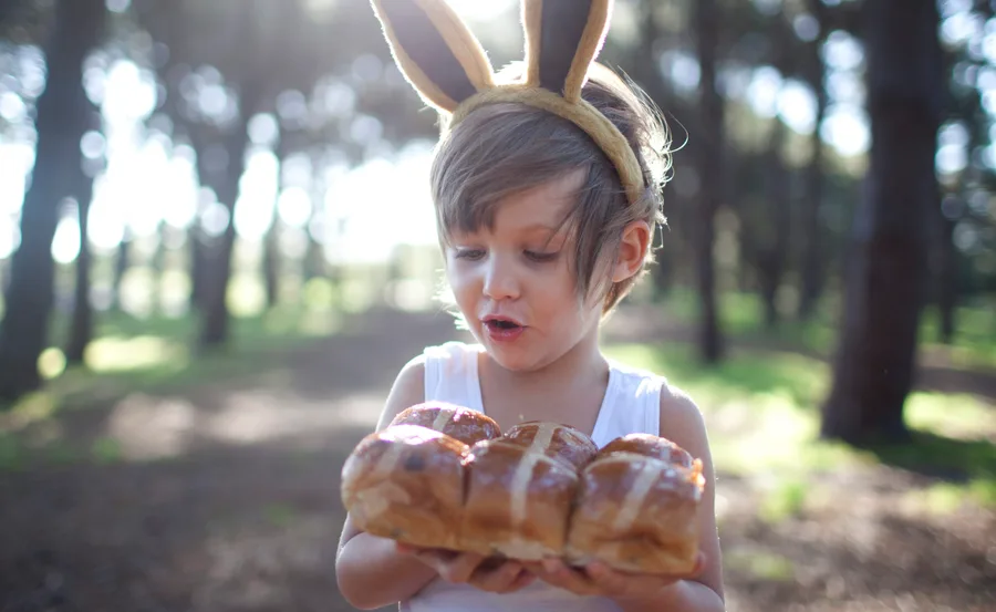 Child with bunny ears holding hot cross buns in a sunlit forest setting.