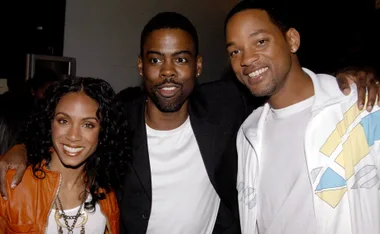 Three people smiling, with one in a dark blazer and the others in light-colored outfits, posing for a photo indoors.
