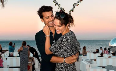 A couple smiling and embracing at a beachside event during sunset, surrounded by people and white decor.