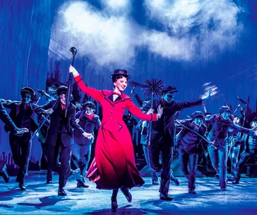 Mary Poppins in a red coat and hat leading a dance number with chimney sweeps on stage, blue background.