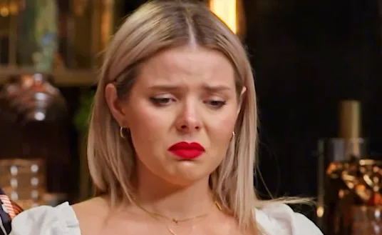 A person with blonde hair looking upset, wearing a white top and red lipstick in a dimly lit setting.