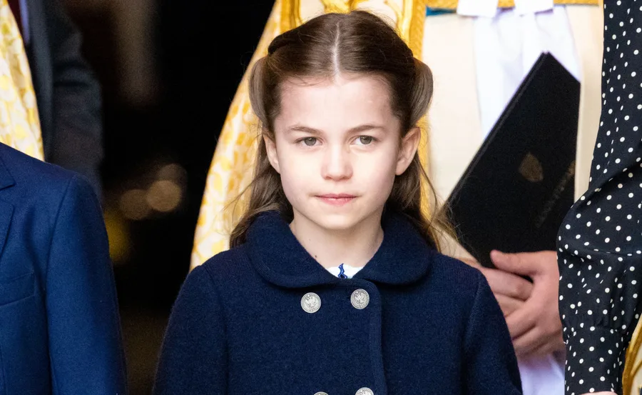 A young girl with long brown hair wearing a blue coat, standing in front of a blurred background.