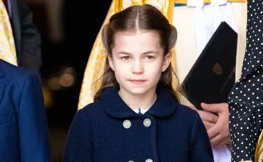 A young girl with long brown hair wearing a blue coat, standing in front of a blurred background.