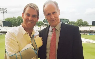 Two men smiling at a cricket stadium, one with an arm cast.