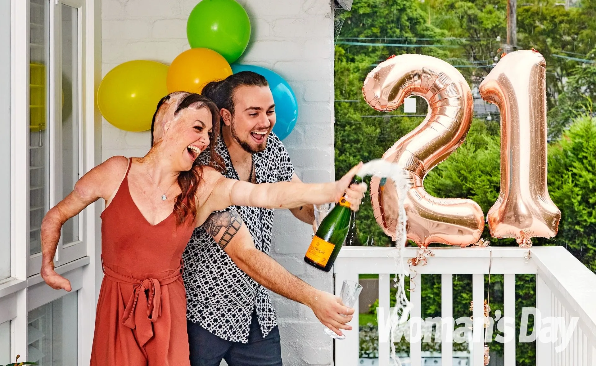 Woman celebrating 21st birthday with a champagne bottle, with big "21" balloons and colorful decorations in the background.