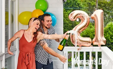 Woman celebrating 21st birthday with a champagne bottle, with big "21" balloons and colorful decorations in the background.