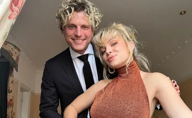 A couple posing indoors, one in a suit, the other in a sparkling dress, both smiling at the camera.