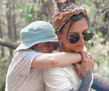 Mother carries her son on her back in a forest, both smiling. She wears sunglasses, a headband, and has tattoos.