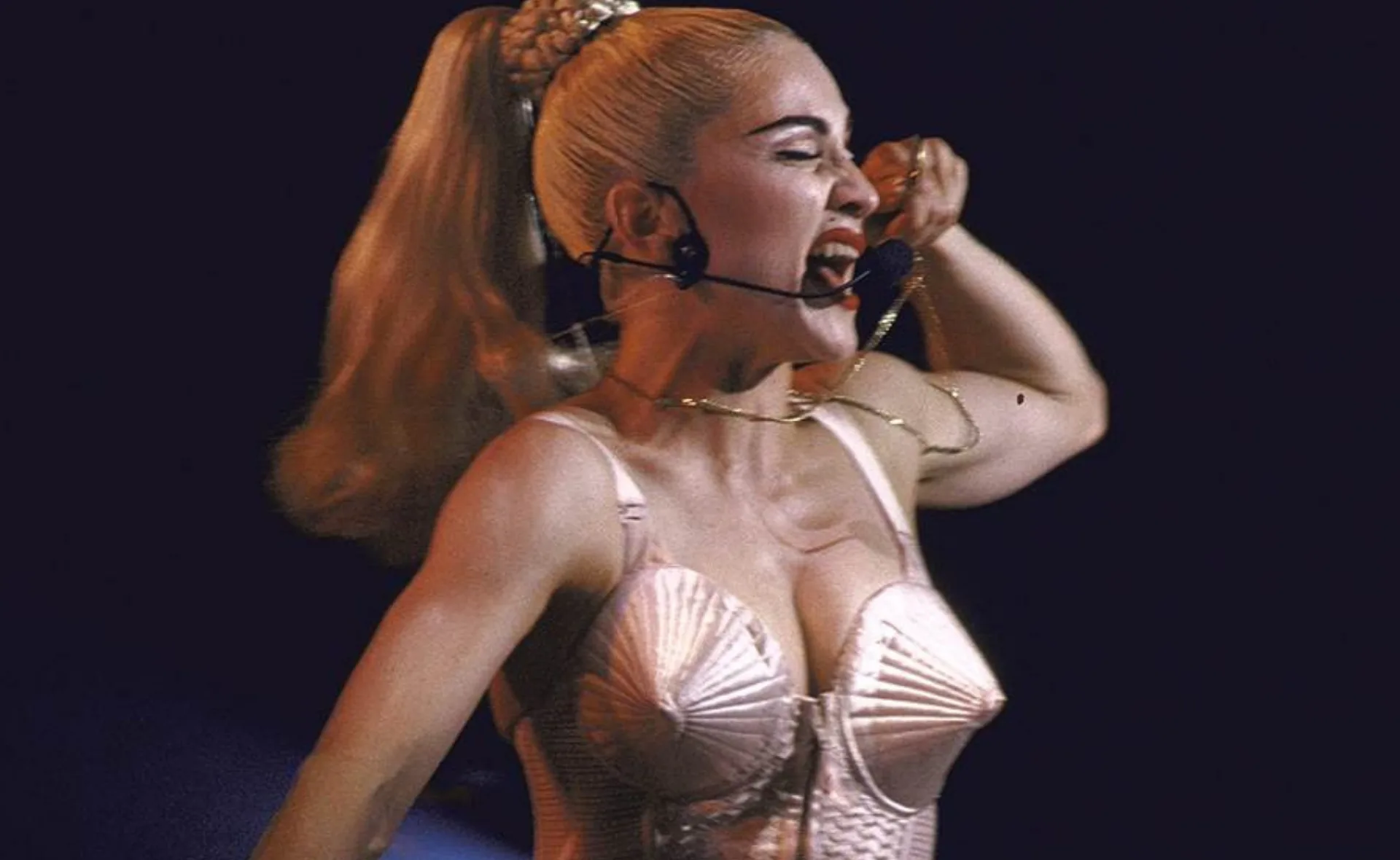 Performer in a conical bra top and headset, energetically singing on stage.