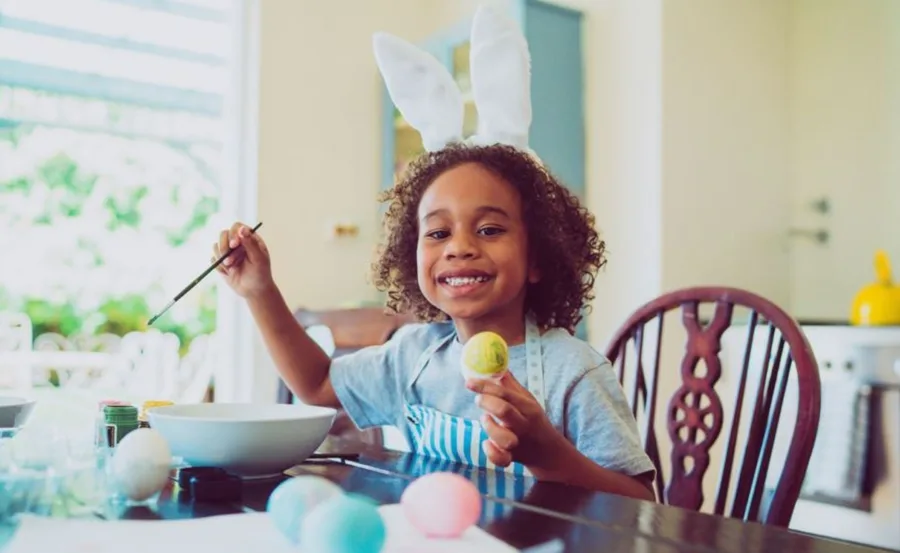 Child with bunny ears painting Easter eggs at a table, smiling and holding a yellow egg.