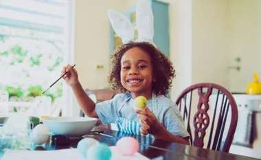 Child with bunny ears painting Easter eggs at a table, smiling and holding a yellow egg.