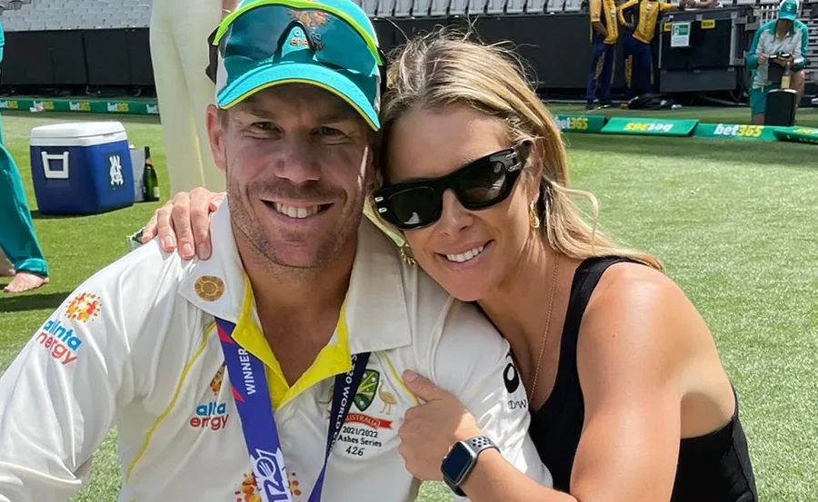 A cricketer in an Australia uniform smiles with a woman in sunglasses at a sports field.