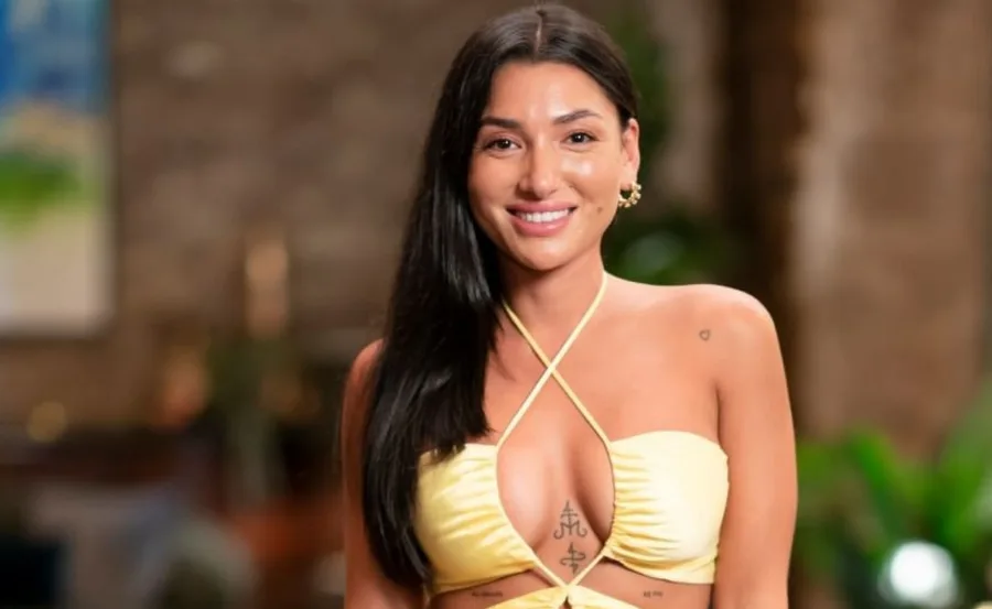 A woman with long dark hair, smiling, wearing a yellow top with tattoos visible on her chest in a blurred background setting.