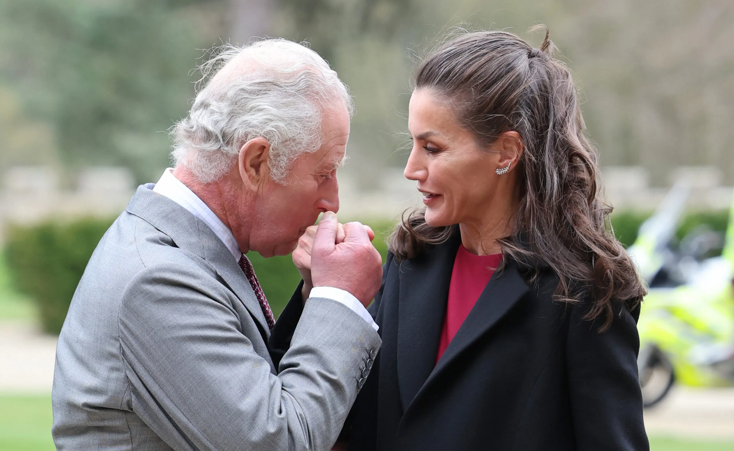 Man in suit kisses woman's hand outdoors, both smiling.