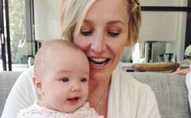 A smiling woman holding a baby indoors, sitting on a sofa with a modern kitchen in the background.