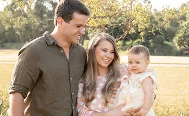 A smiling couple holds a baby girl outdoors, surrounded by nature under a sunny sky.