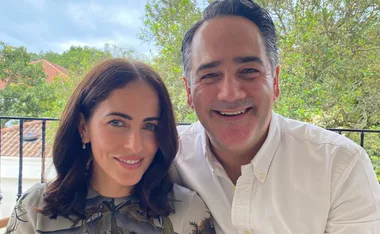 A smiling couple poses together outdoors with greenery in the background.