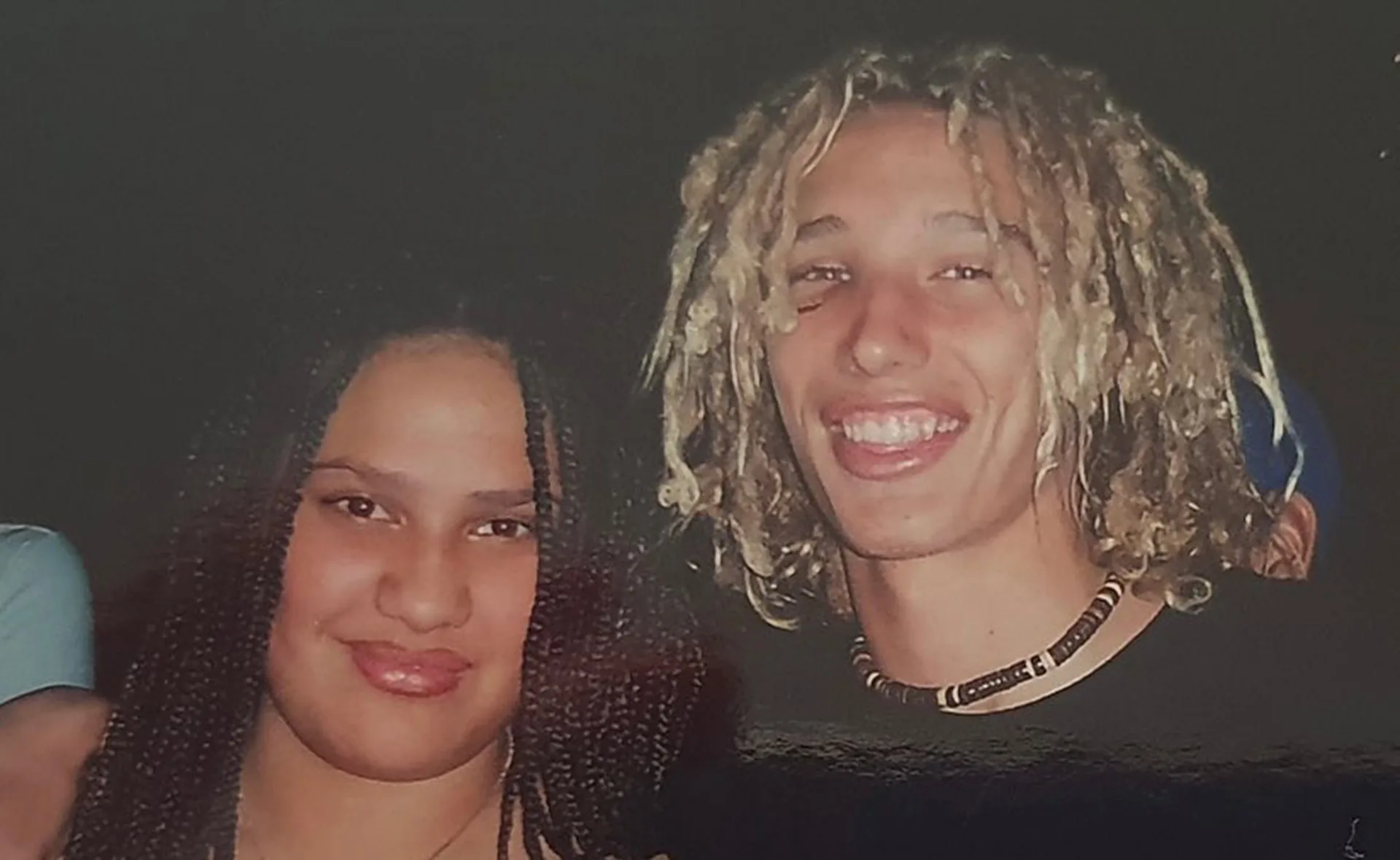 A young man with blond dreadlocks and a necklace smiles next to a woman with braided hair.