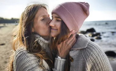 A mother kisses her smiling daughter on a beach, both wearing cozy sweaters, creating a warm, affectionate moment.