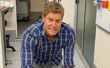 Man in a blue plaid shirt kneeling indoors with a humorous expression.
