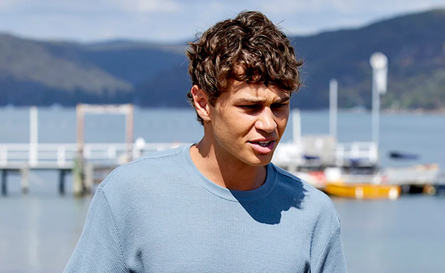 Young man in a blue shirt outdoors by a waterfront with docks and hills in the background.