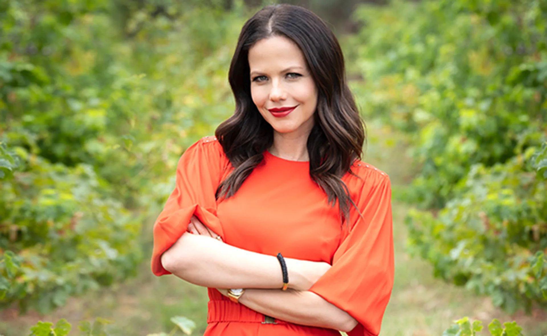 Tammin Sursok in an orange dress with arms crossed, standing in a lush green garden setting.