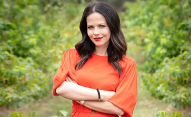 Tammin Sursok in an orange dress with arms crossed, standing in a lush green garden setting.