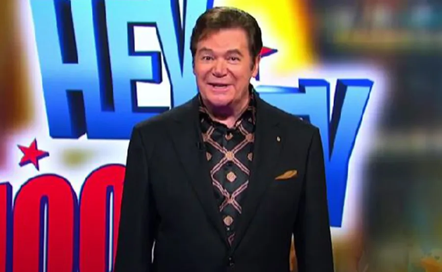 Host in a patterned shirt stands in front of "Hey Hey It's Saturday" backdrop, smiling.