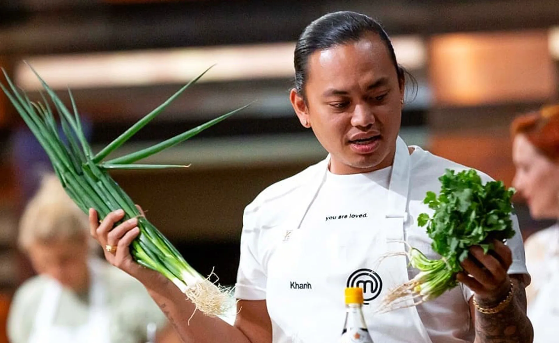 Chef holds fresh green onions and cilantro in a cooking competition kitchen, wearing a white apron.