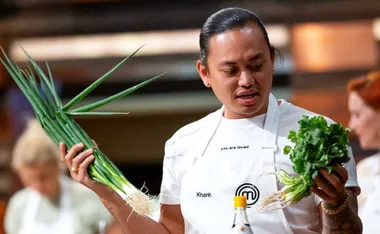Chef holds fresh green onions and cilantro in a cooking competition kitchen, wearing a white apron.