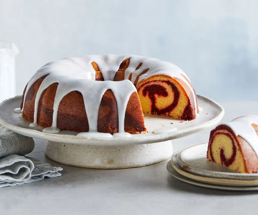 Red velvet marble bundt cake with white icing, partially sliced to show the red swirl inside, on a cake stand.