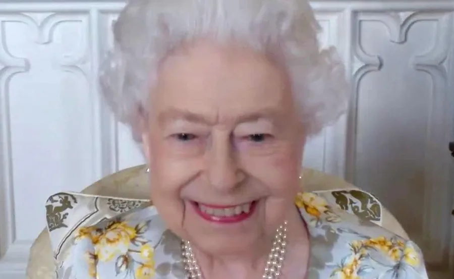 A smiling elderly woman with white hair, wearing a floral dress and pearl necklace, seated indoors.
