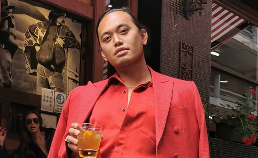 Person in a red suit holding a drink, standing outside a building with a vintage poster in the background.