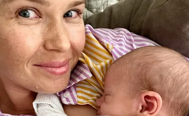 A woman in a striped shirt smiles while holding a sleeping baby close to her chest.