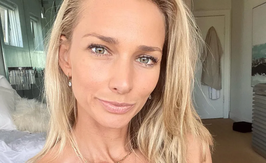 A woman with blonde hair smiling indoors, wearing earrings and a necklace.