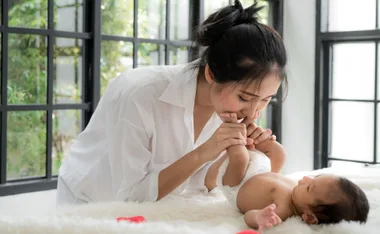 Mother in white shirt kisses baby's feet, both on a bed near large windows.