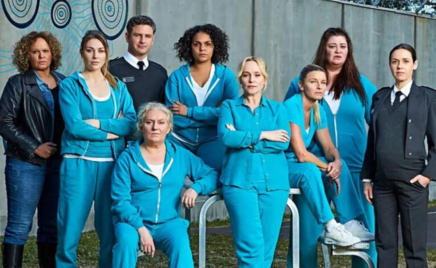 "Cast of 'Wentworth' series in teal uniforms, posed against a wall with circular designs, including uniformed officers."