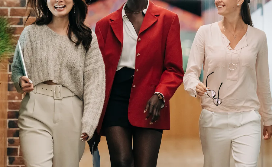 Women walking confidently in stylish office attire, smiling and holding documents and glasses.