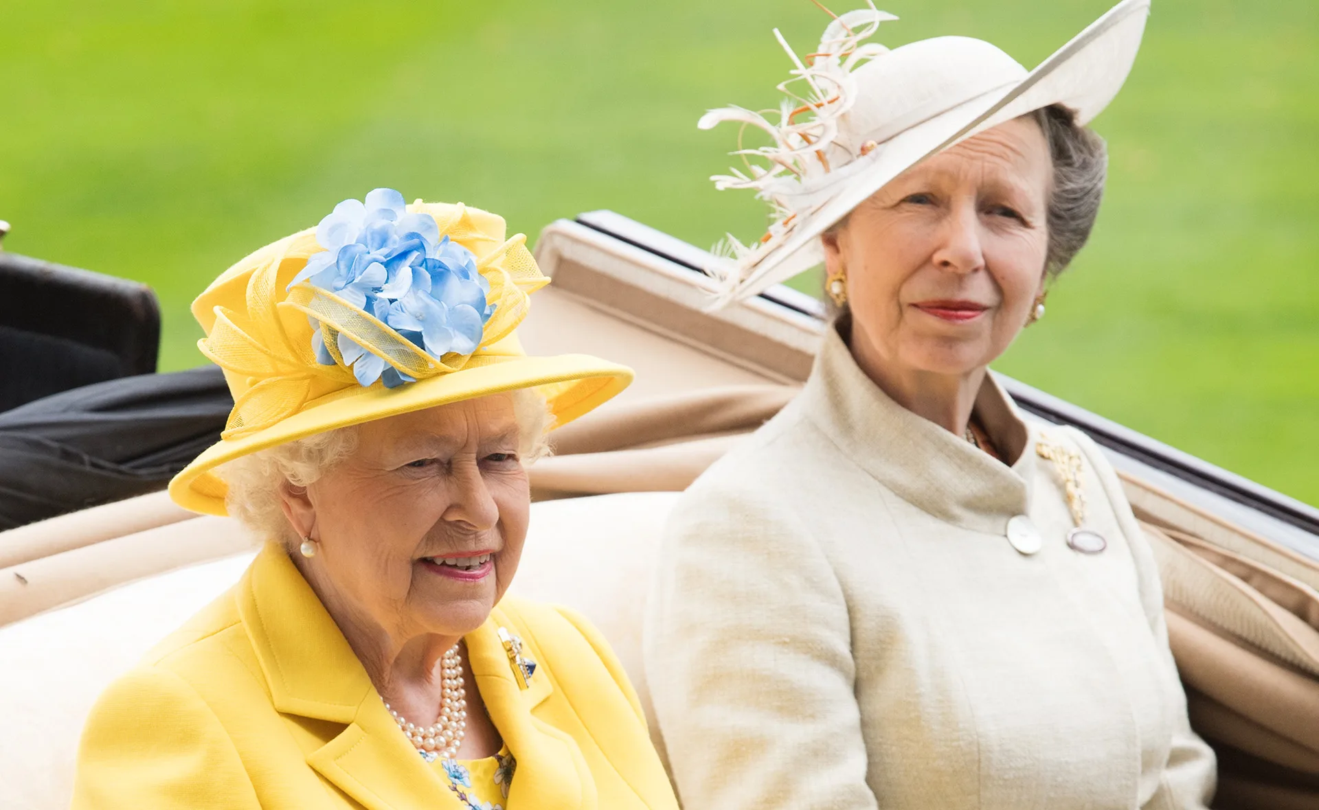 Two women sitting outdoors in elegant attire with decorative hats, smiling and looking ahead.