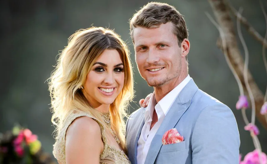 A smiling woman in a gold dress stands beside a man in a light blue suit, with a blurred natural background.