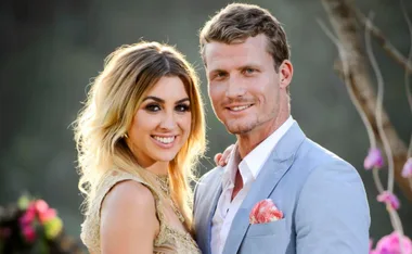 A smiling woman in a gold dress stands beside a man in a light blue suit, with a blurred natural background.