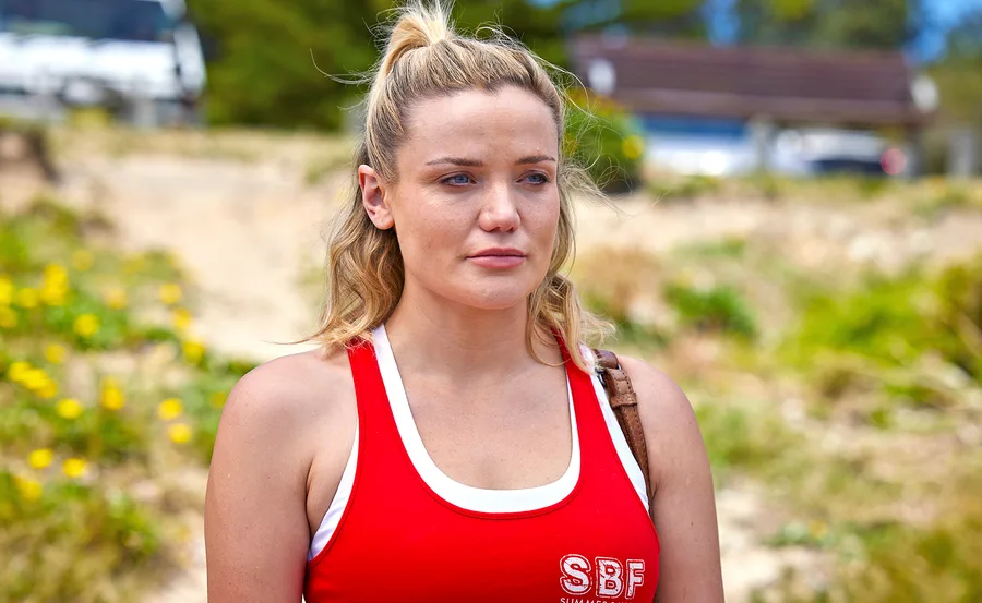 Character from Home and Away in a red tank top with "SBF" printed, standing outdoors with a serious expression.