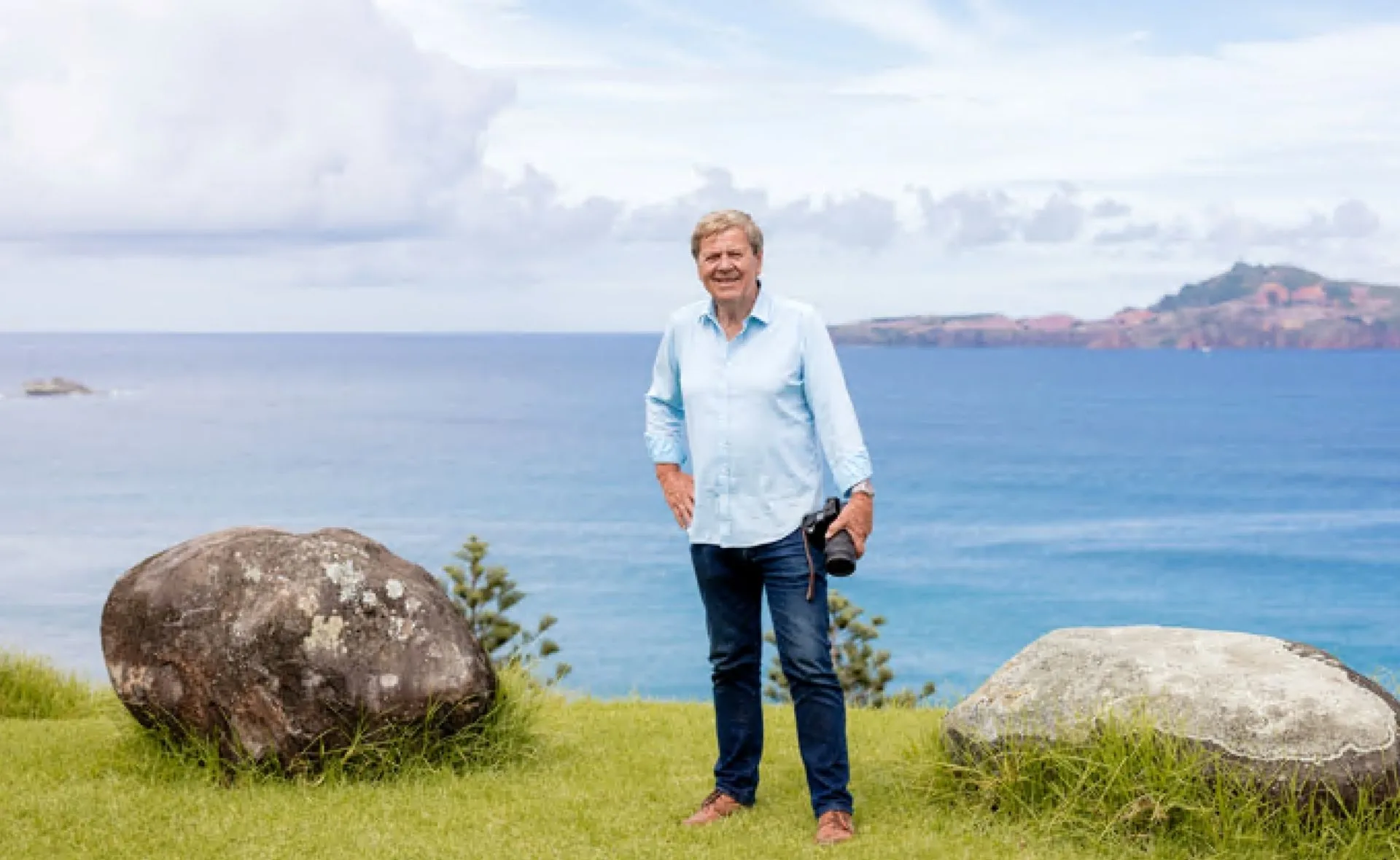 Man in a blue shirt standing on grass with rocks, holding a camera near a scenic ocean view.