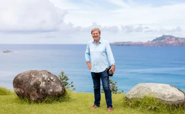 Man in a blue shirt standing on grass with rocks, holding a camera near a scenic ocean view.