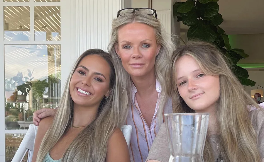 Three women smiling at a table indoors, with plants and a glass jug in the foreground.