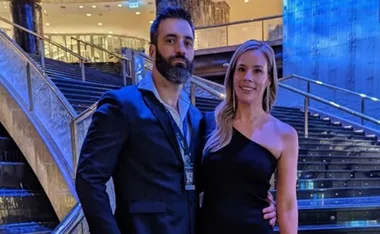 A couple posing in front of a modern staircase, both dressed in formal evening attire.