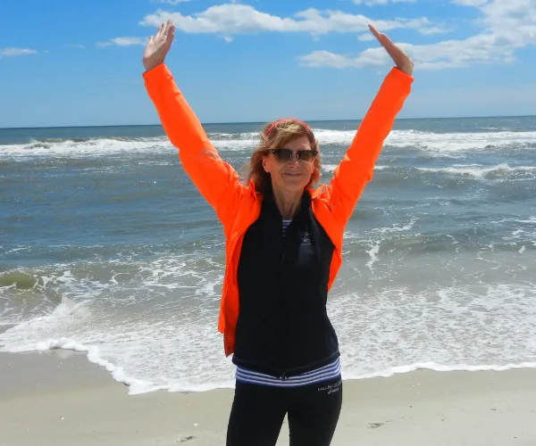 Person in an orange jacket stands on a beach with arms raised, ocean waves behind them.